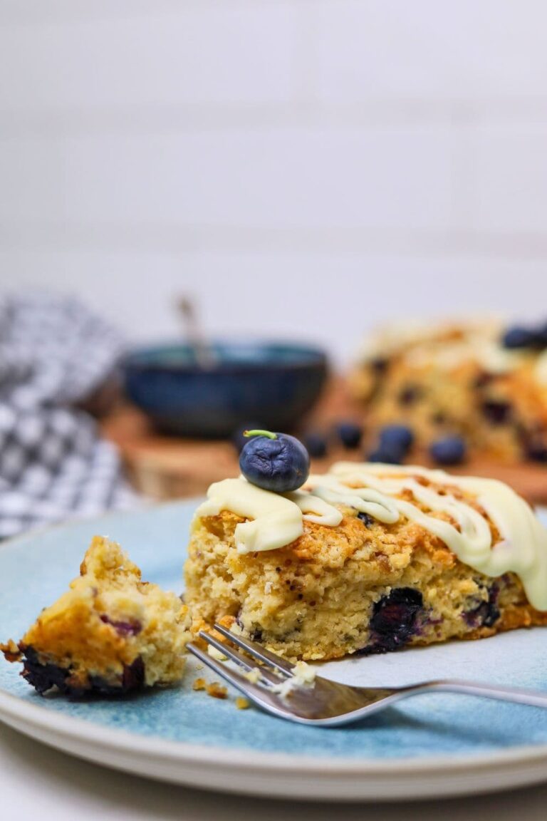 Close up photo of a sourdough blueberry scone drizzled in white chocolate. Part of the scone has been broken away with a fork.