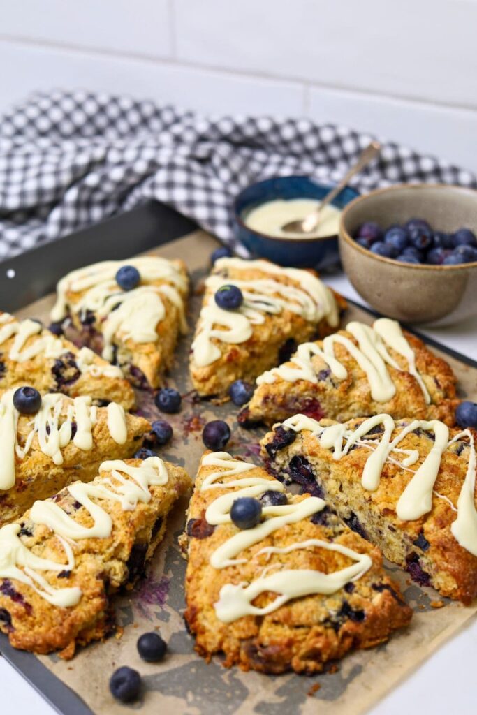 8 wedges of sourdough blueberry scones drizzled in white chocolate on a baking tray. There is a bowl of blueberries to the right of the photo.