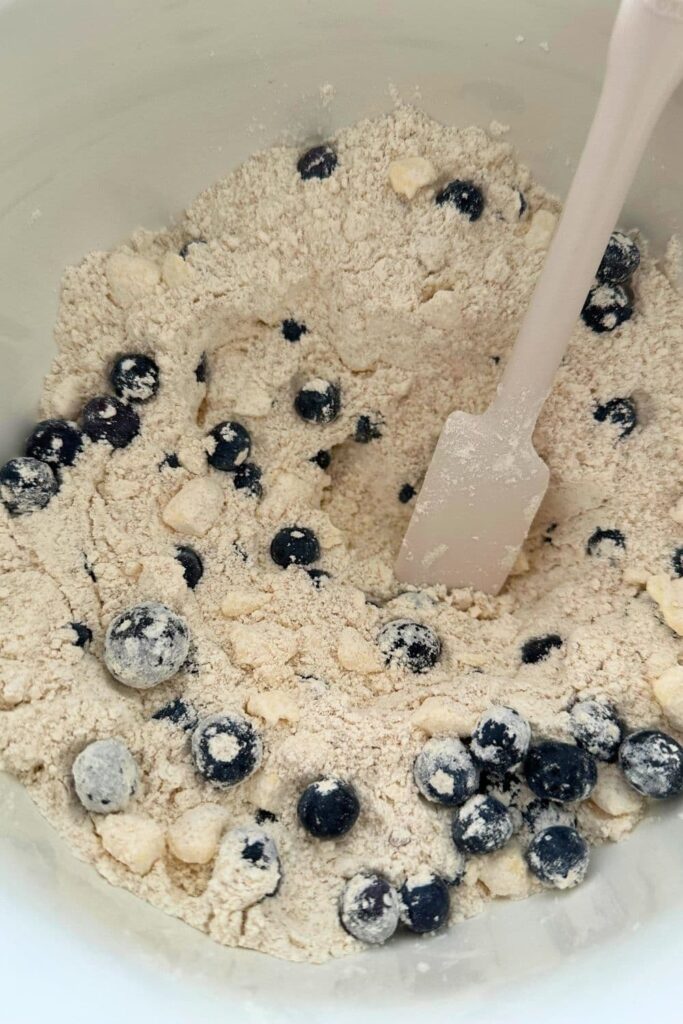 A bowl of flour with butter rubbed into it. There are blueberries stirred through the flour and a white spatula in the bowl. 