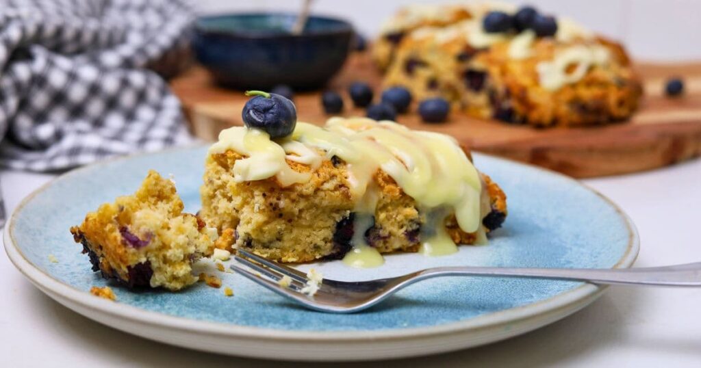 Close up photo of a sourdough blueberry scone drizzled in white chocolate ganache. Part of the scone has been broken away with a fork.