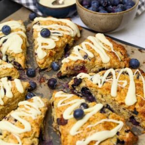 A tray of sourdough blueberry scones drizzled in white chocolate and surrounded by fresh blueberries.