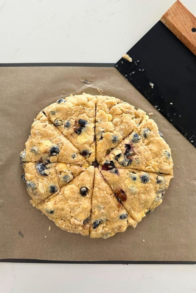 A baking tray with a round disc of sourdough blueberry scone dough on it. The dough has been cut into 8 wedges.