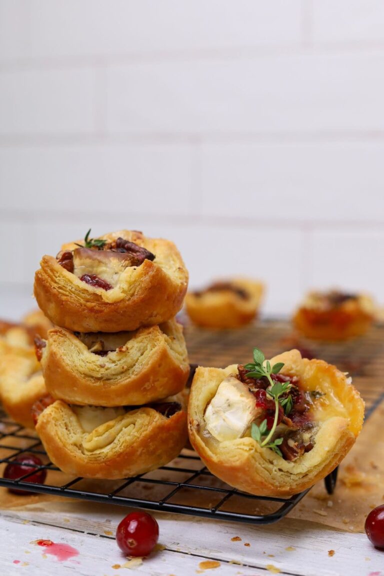 A stack of three sourdough brie and cranberry puff pastry tarts sitting on a wire cooling rack. There is a also a single tart sitting next to the stack that has been garnished with fresh thyme.