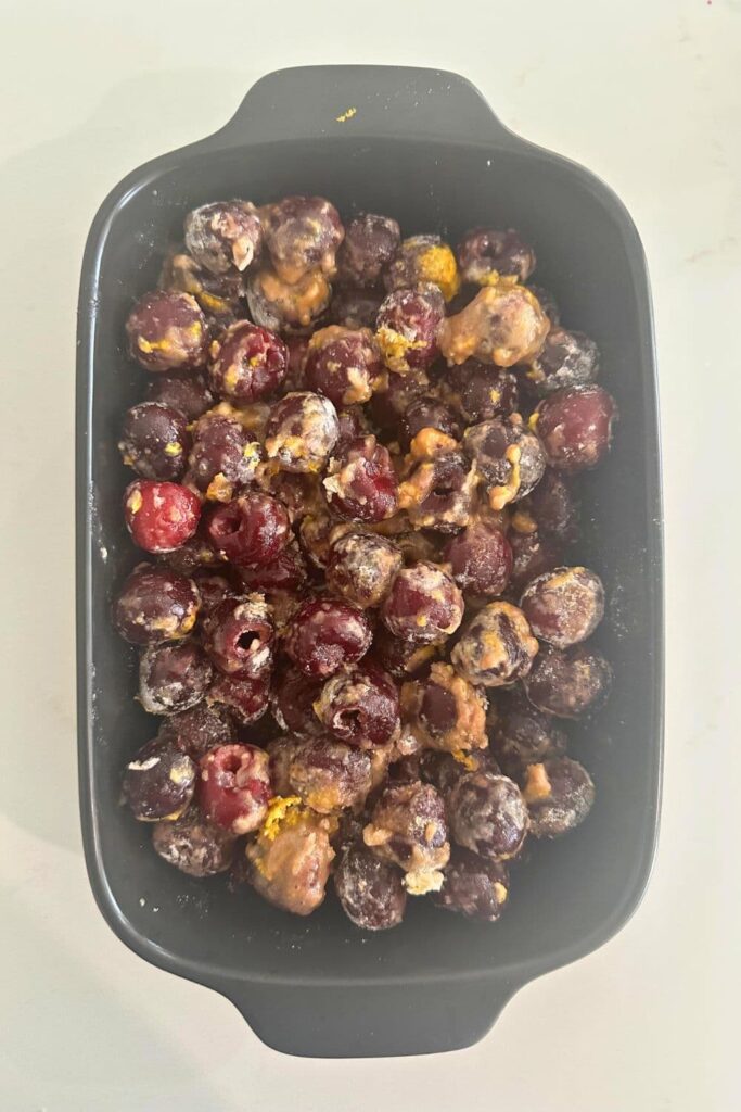 A black baking dish filled with cherries coated in flour, sugar and orange zest.