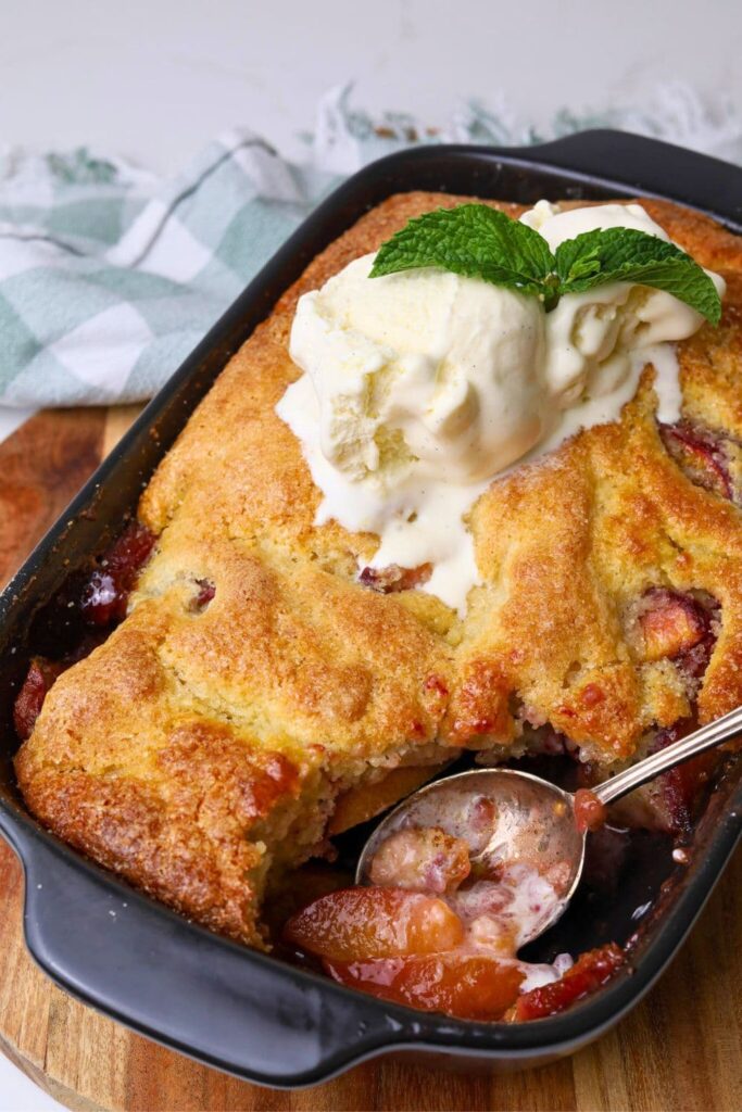 A black baking dish filled with sourdough peach cobbler topped with vanilla ice cream. There is a vintage spoon that has taken a scoop out of the dish so you can see the juicy peaches underneath.