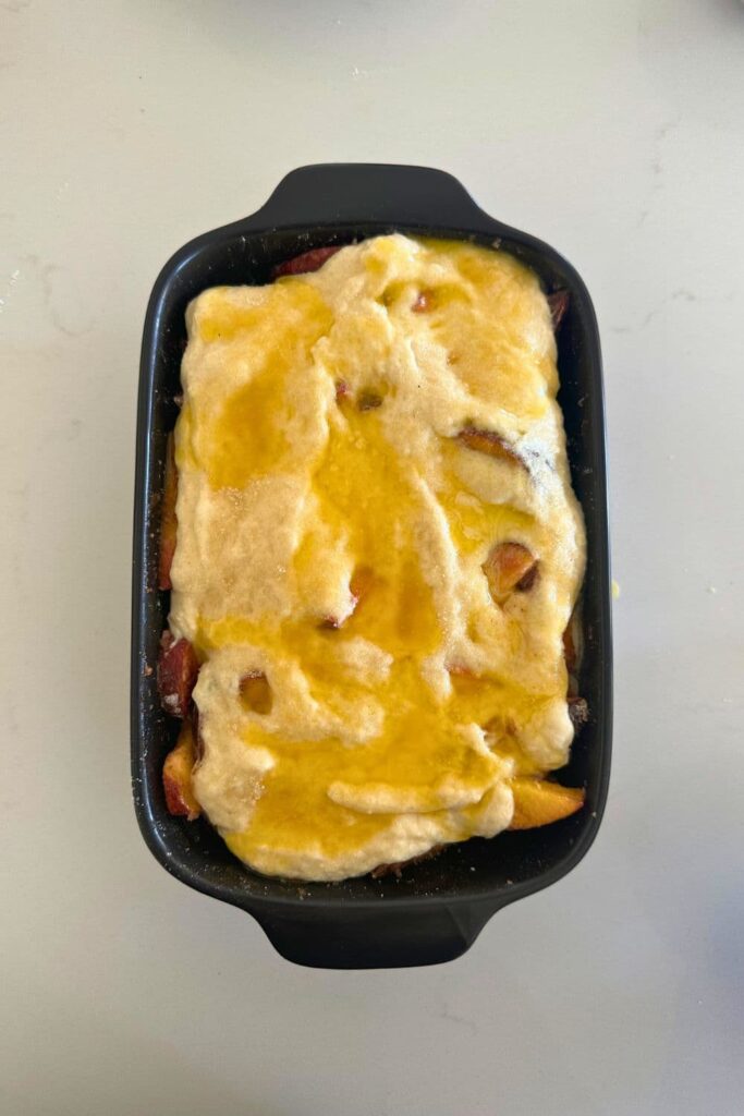 A black casserole dish filled with peach cobbler topped with a sourdough sponge batter that has butter poured over it.