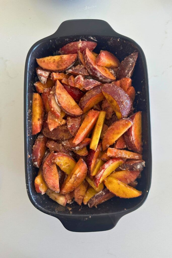 A black baking dish filled with chopped peaches coated in brown sugar and lemon juice.