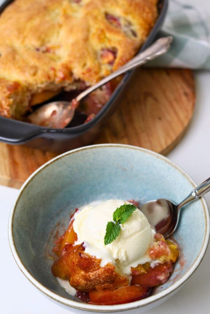 A light blue bowl containing a generous scoop of sourdough peach cobbler topped with vanilla ice cream. You can see the rest of the sourdough peach cobbler sitting in the background of the photo.