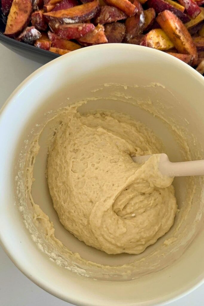 A cream colored mixing bowl filled with a sponge cobbler batter being mixed with a white rubber spatula. You can see a peak of the peach cobbler filling in the background of the photo.