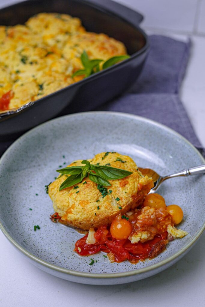 A shallow bowl with a single serve of sourdough tomato cobbler served out. You can see the red and yellow tomatoes of the filling oozing out from underneath the biscuit topping. You can see the rest of the sourdough tomato cobbler in the black baking dish in the background.