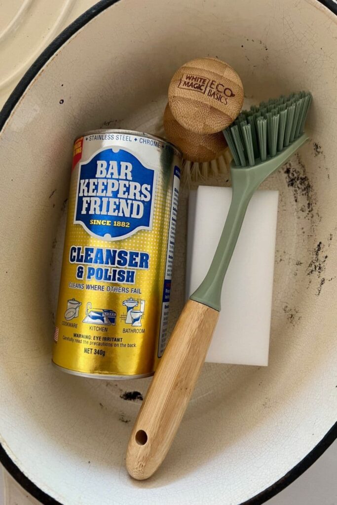 A photo taken from above showing the inside of a Dutch Oven. Inside the Dutch Oven is a jar of Bar Keepers Friend as well as 2 scrubbing brushes and a magic eraser.