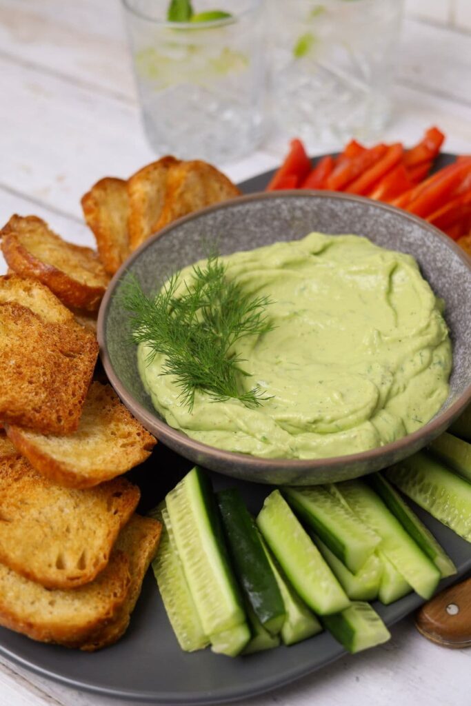 A dish of smooth avocado dip garnished with fresh dill and surrounded by sourdough crostini and fresh vegetable sticks.