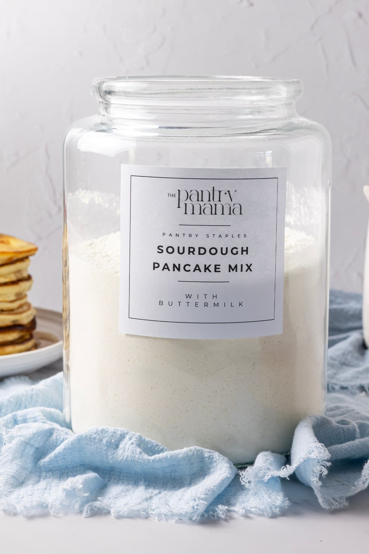 A large glass jar filled about three quarters full with homemade sourdough pancake mix. There is a pale blue linen cloth under the jar and a black and white label on the front that says Sourdough Pancake Mix.