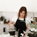 A photo of a woman (Kate from The Pantry Mama) pouring water into a glass jar that is sitting on a scale. She is feeding her sourdough starter.