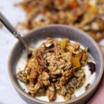 A bowl of sourdough granola and yoghurt being held in a hand. There is a spoon in the bowl and there is more sourdough granola that has been spilled around the bowl.