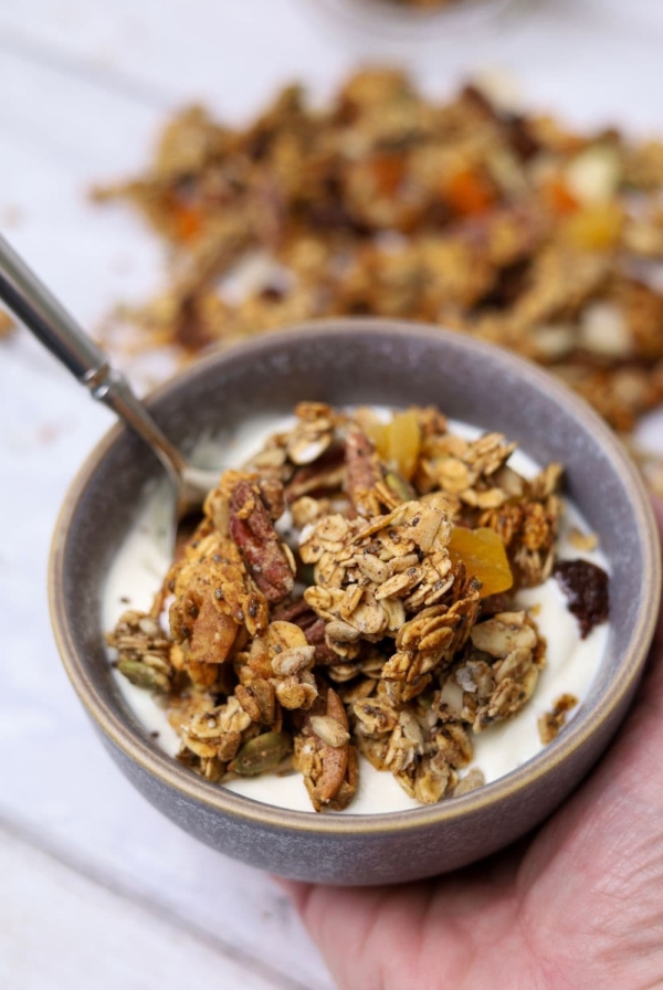 A bowl of sourdough granola and yoghurt being held in a hand. There is a spoon in the bowl and there is more sourdough granola that has been spilled around the bowl.