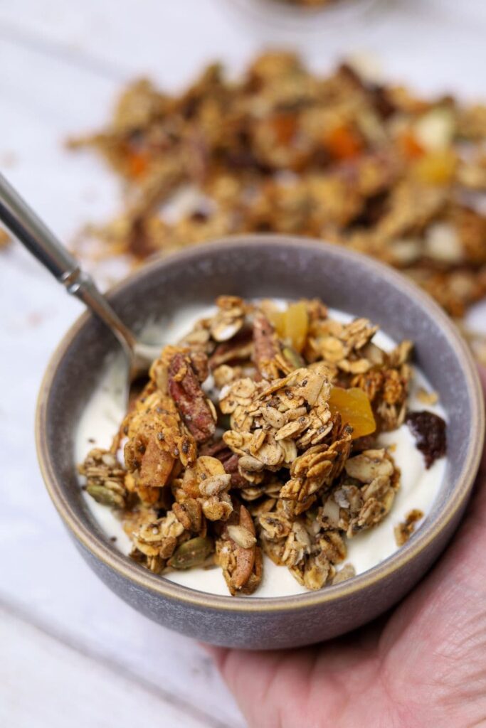 A bowl of sourdough granola and yoghurt being held in a hand. There is a spoon in the bowl and there is more sourdough granola that has been spilled around the bowl.