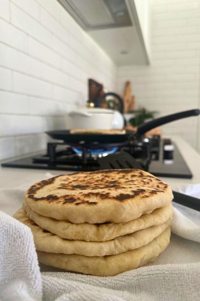 A stack of sourdough pita breads sitting on a dish towel. In the background you can see a skillet on a gas cook top where the sourdough pita breads are being cooked.