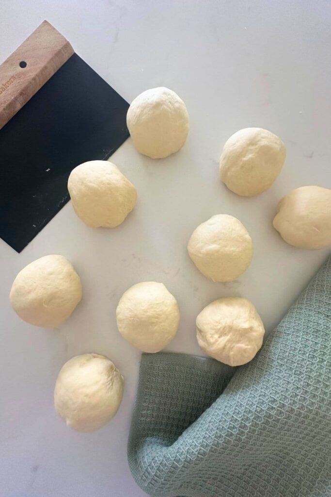 Sourdough pita bread dough that has been divided into small portions. You can also see a black metal dough scraper with a wooden handle and a green dish towel.