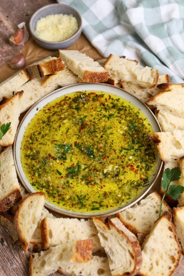 A shallow dish of bread dipping oil filled with herbs and parmesan cheese surrounded by pieces of sourdough bread.