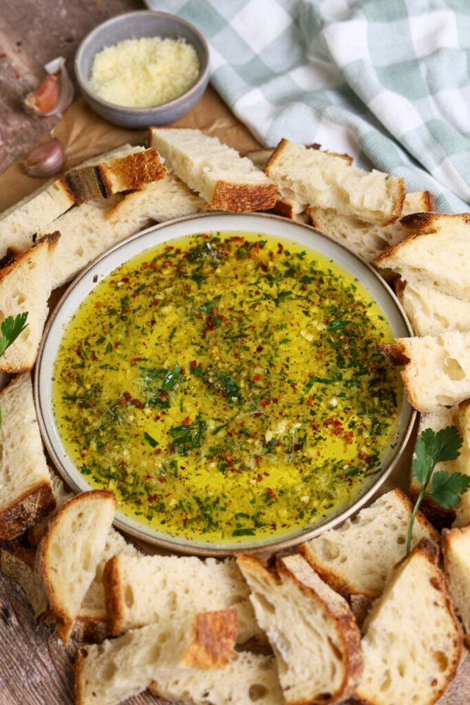 A shallow dish of bread dipping oil filled with herbs and parmesan cheese surrounded by pieces of sourdough bread.