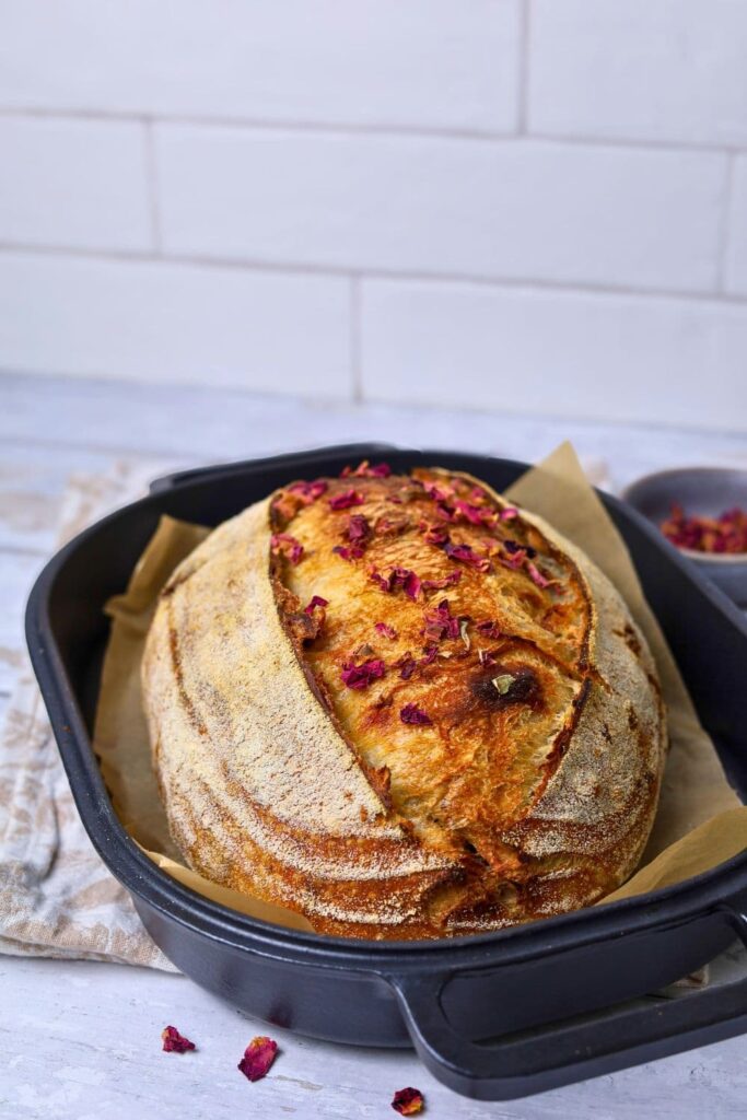 A loaf of sourdough bread topped with dried rose petals. The sourdough bread is infused with dried rose petals, cinnamon and white chocolate and is sitting in a cast iron bread pan.