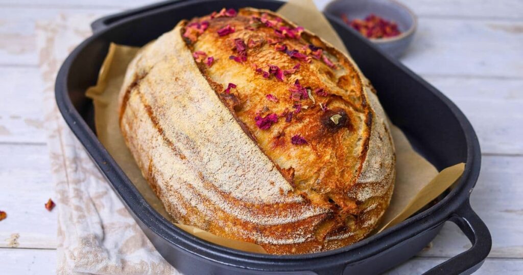 A horizontal image of rose petal sourdough bread that has been baked. It is sitting in the cast iron bread pan it was baked in and has been sprinkled with dried rose petals.