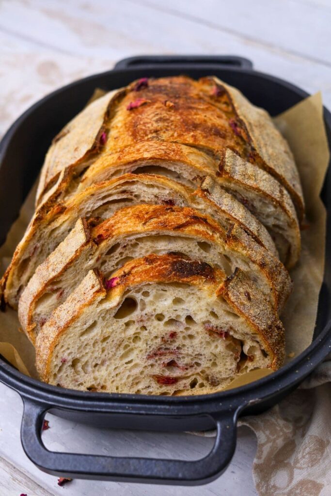 A loaf of sourdough bread infused with dried rose petals, cinnamon and white chocolate that has been sliced and placed into a cast iron bread pan. You can see the pink rose petals in the crumb of the sourdough.