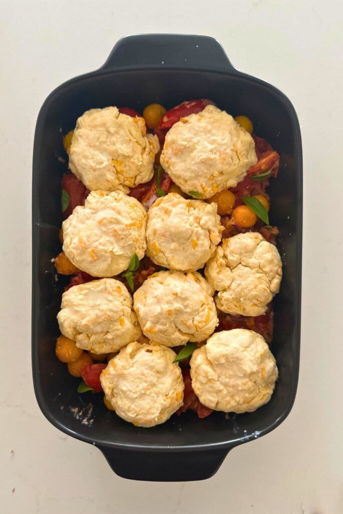 A sourdough tomato cobbler that has been topped with sourdough cheddar biscuits that is ready to be baked in the oven.
