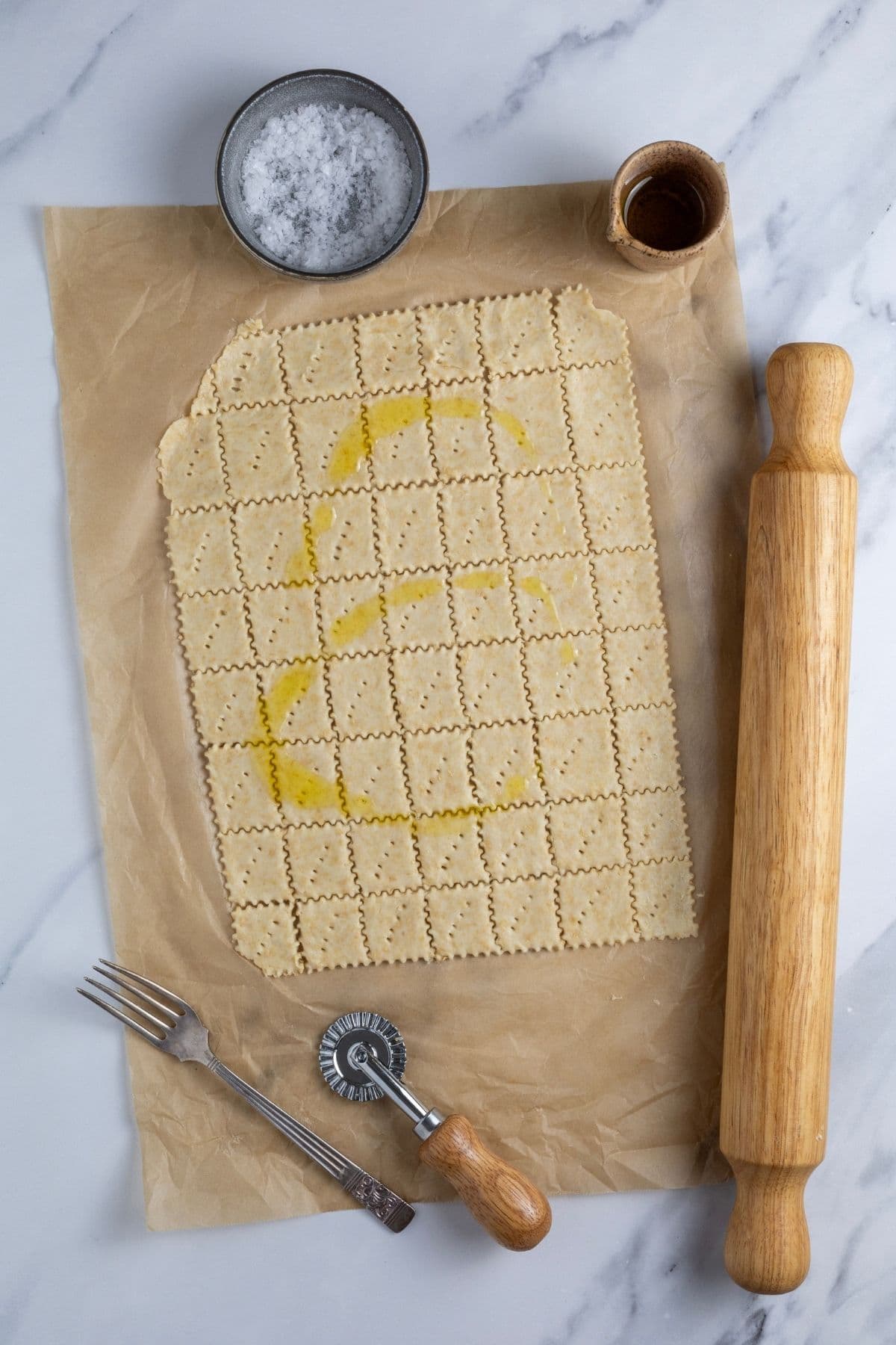 Whole wheat sourdough cracker dough cut into squares and drizzled with olive oil. There is a vintage fork, ravioli cutter, rolling pin, small dish of salt and rolling pin surrounding the cut crackers.