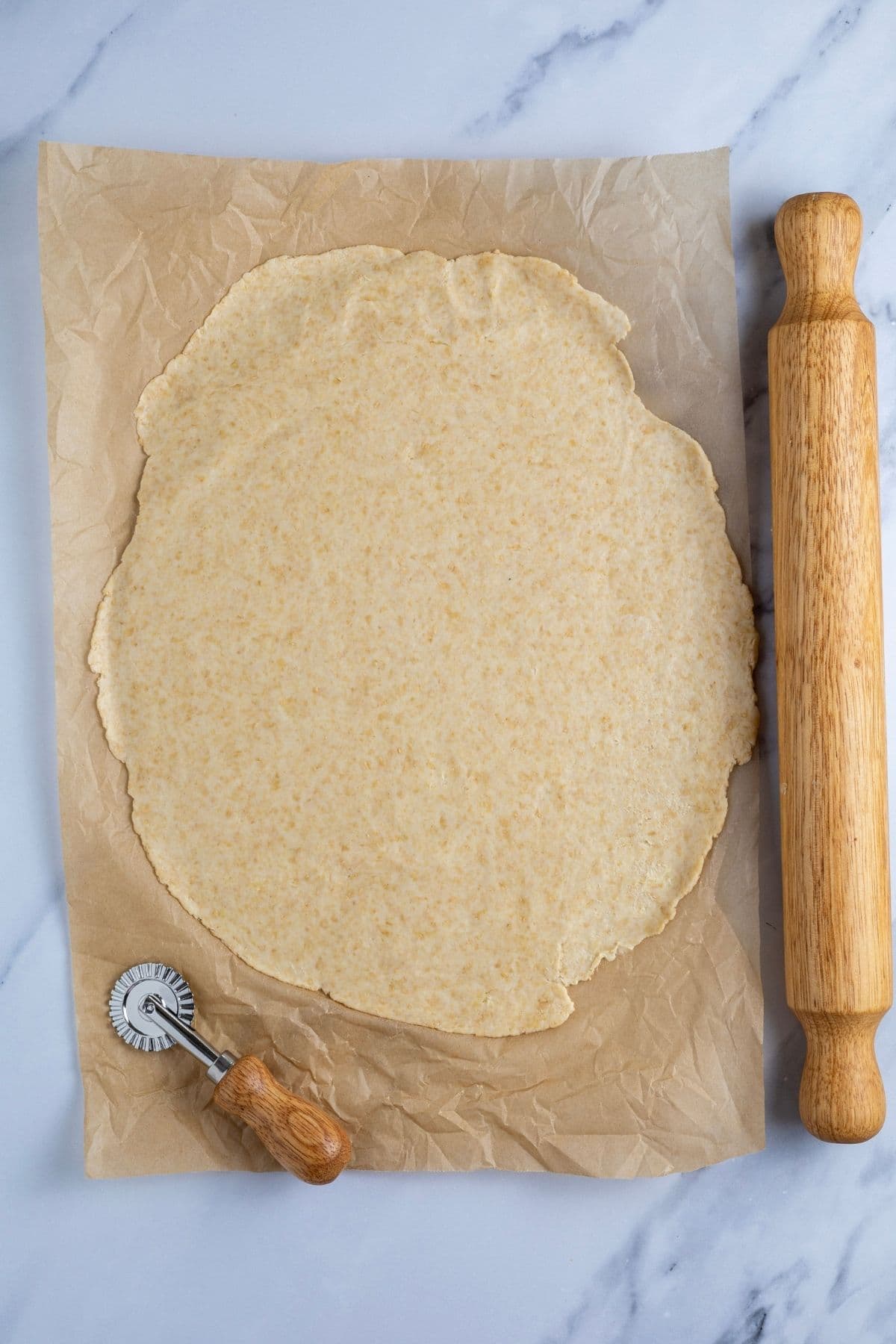 A sheet of whole wheat sourdough cracker dough rolled out really thin on a piece of parchment paper. There is a rolling pin and a small ravioli cutter in the photo too.