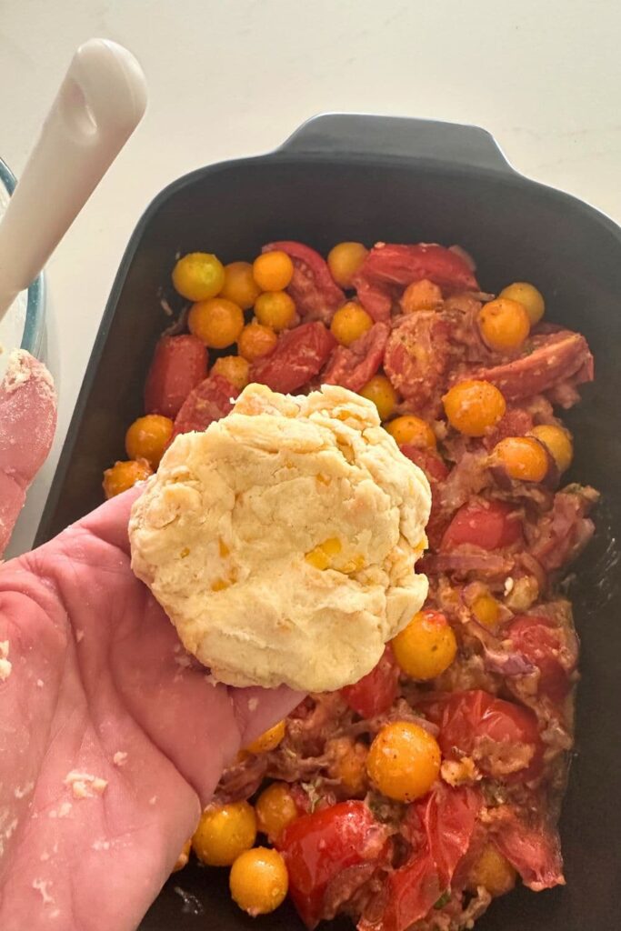 A photo of the base of a sourdough tomato cobbler in a black baking dish. There a hand holding the sourdough cheddar biscuit topping as it's being added to the base.