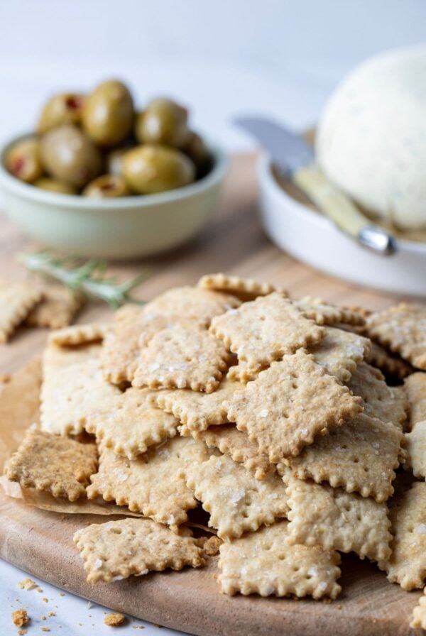 A generous stack of whole wheat sourdough crackers on a wooden board. In the background there is a small bowl of olives and some homemade Boursin cheese.