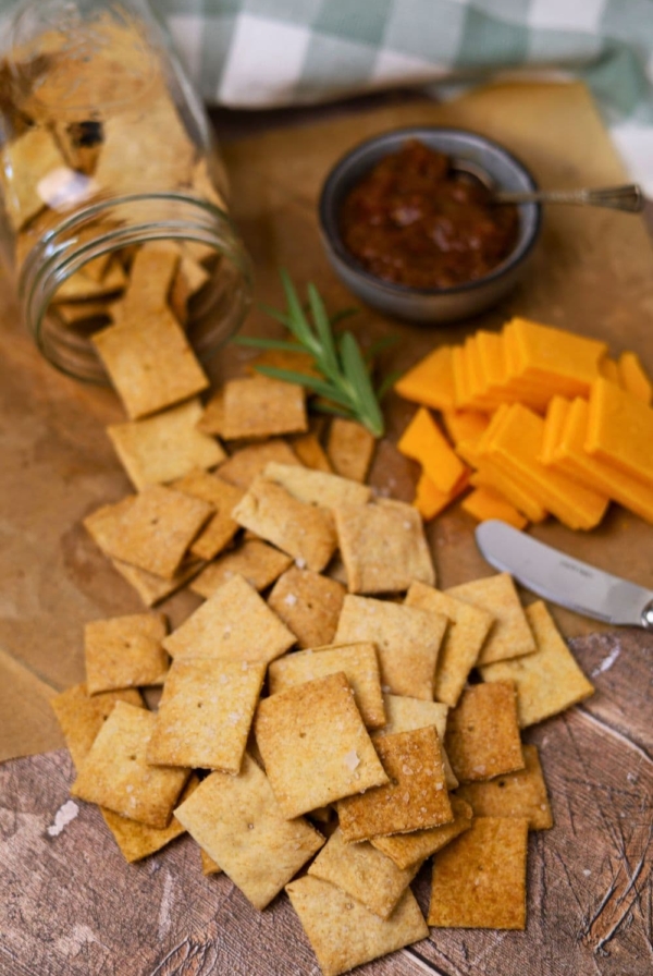 A display of sourdough whole wheat crackers that have been tipped out of a jar onto a counter top. There is also some cheese and tomato relish in the photo and it has been garnished with some fresh rosemary.
