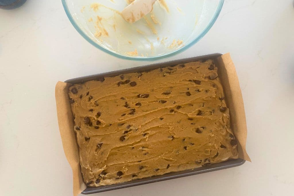 A tray of sourdough chocolate chip cookie bars ready to be baked in the oven. There is also an empty bowl at the top of the photo as well.