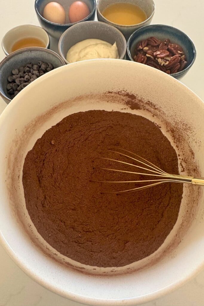 A cream bowl filled with the dry ingredients necessary to make spiced sourdough brownies. The other ingredients are in the background of the photo.