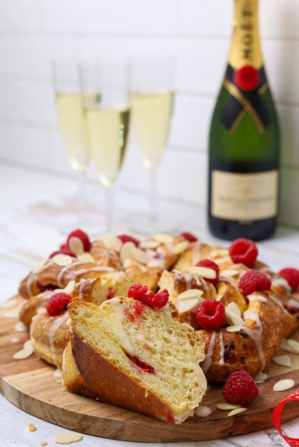 A sourdough brioche braided loaf decorated with flaked almonds and fresh raspberries displayed on a wooden board. There are 3 glasses of champagne and a bottle of Moet in the background of the photo.