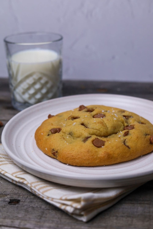 Single serve sourdough chocolate chip cookie sitting on a white plate. There is a glass of milk in the background.