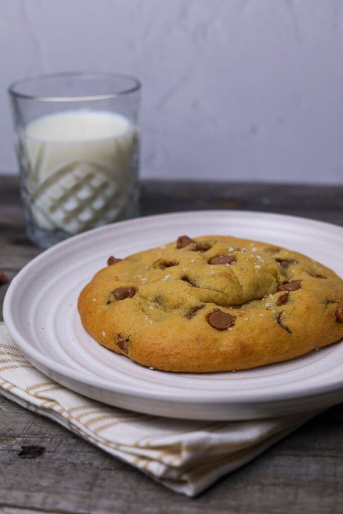 Single serve sourdough chocolate chip cookie sitting on a white plate. There is a glass of milk in the background.