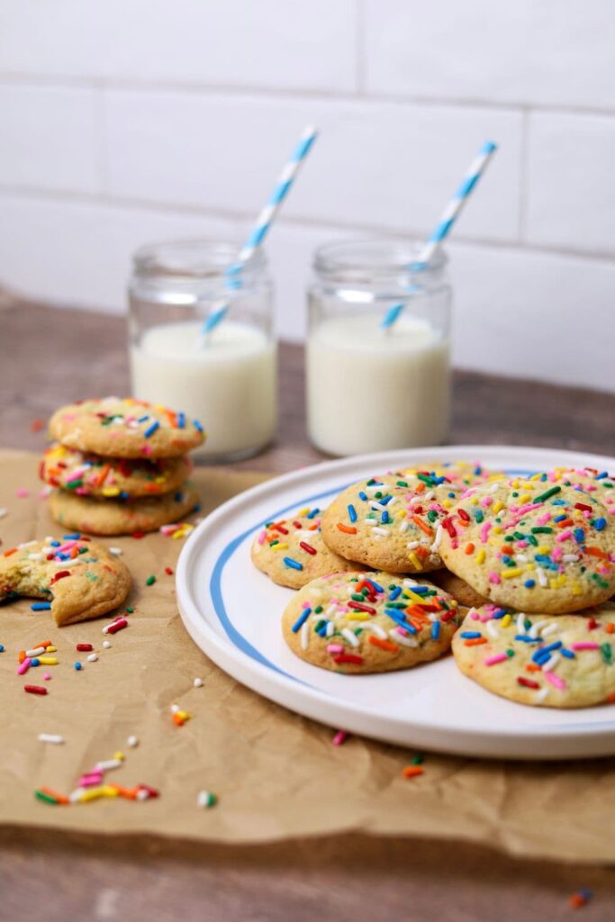 A blue edged plate stacked with colorful sourdough funfetti cookies sits to the left of the photo. There are two glasses of milk with blue striped straws in the background, as well as some cookies with bites taken out of them.