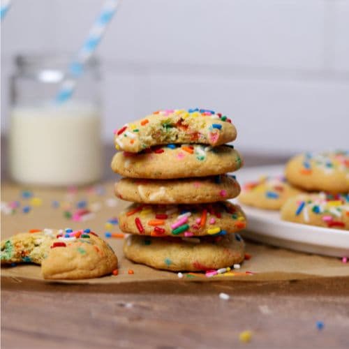 A stack of 5 sourdough funfetti cookies. The top cookie has a bite taken out of it, as does the cookie sitting next to the stack. There is a glass of milk with a blue straw in the background of the photo.