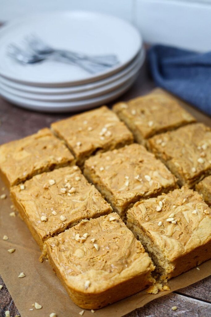 9 sourdough peanut butter squares topped with crushed peanuts and set in front of a stack of white plates topped with cake forks.