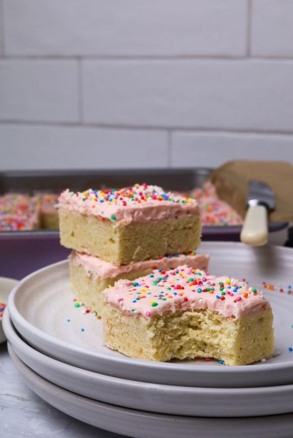 A slice of sourdough sugar cookie bar topped with strawberry frosting and rainbow sprinkles displayed on a white plate. There is a bite taken out of the slice and two other pieces behind it stacked on top of each other.