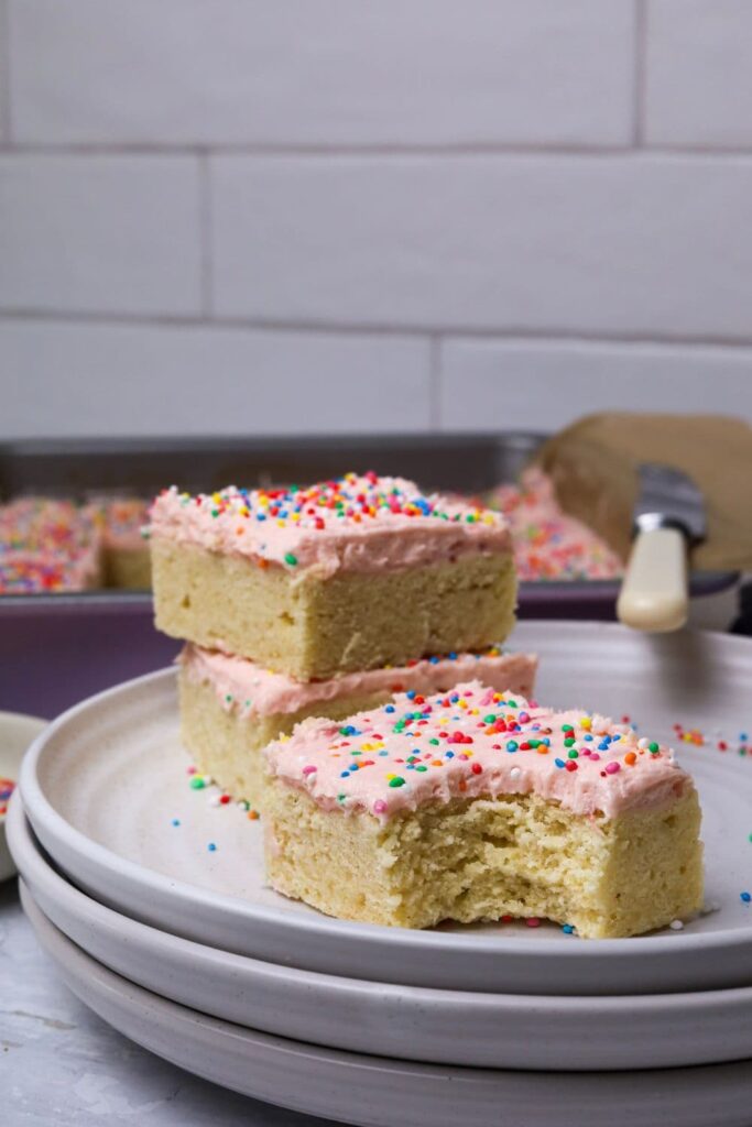 A slice of sourdough sugar cookie bar topped with strawberry frosting and rainbow sprinkles displayed on a white plate. There is a bite taken out of the slice and two other pieces behind it stacked on top of each other.