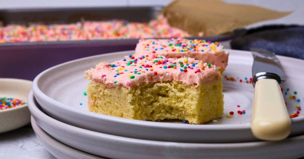 A slice of sourdough sugar cookie bar topped with strawberry frosting and rainbow sprinkles displayed on a white plate. There is a bite taken out of the slice and another piece behind it.