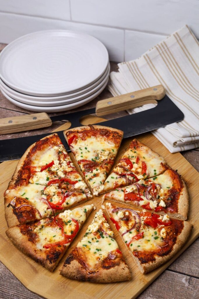 A whole wheat sourdough pizza crust that has been baked and sliced. There is a stack of white plates and a pizza rocker in the background.