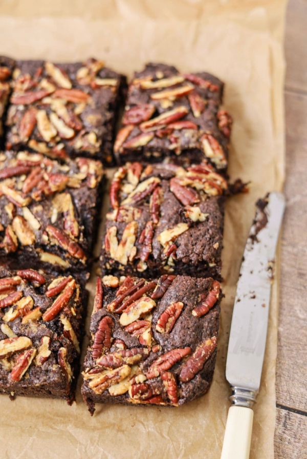 6 squares of spiced sourdough brownies topped with buttered pecans sitting on a sheet of parchment paper. There is a butter knife sitting next to the brownies.