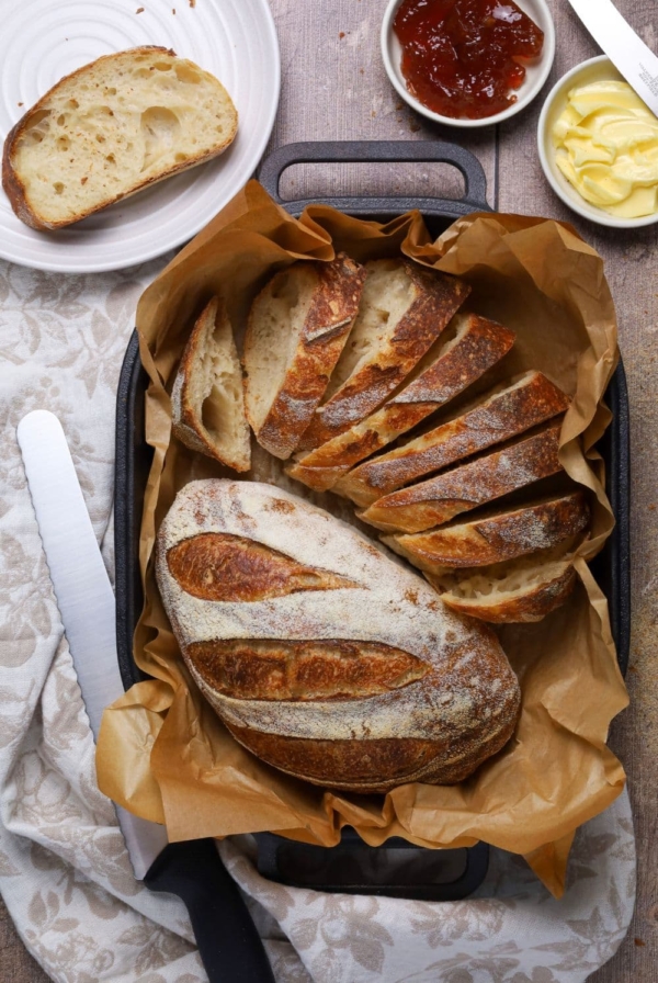An overhead photo showing two small loaves that have been baked in a cast iron bread pan. One the loaves has been sliced up, the other one is whole. You can see one of the slices of bread on a plate to the top left of the photo and two small dishes containing butter and jam to the top right. There is also a black handled bread knife in the photo.