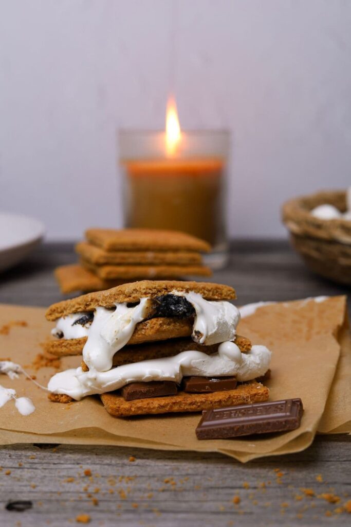 2 s'mores made with sourdough graham crackers. The marshmallow is super melty and oozing out. There is a stack of sourdough graham crackers and a lit candle in the background.