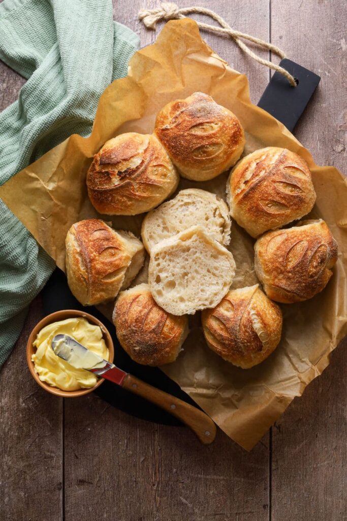 8 crusty sourdough bread rolls arranged in a circle with one of the rolls cut in half in the centre. There is a small dish of butter and a sage green dish towel also displayed in the photo.